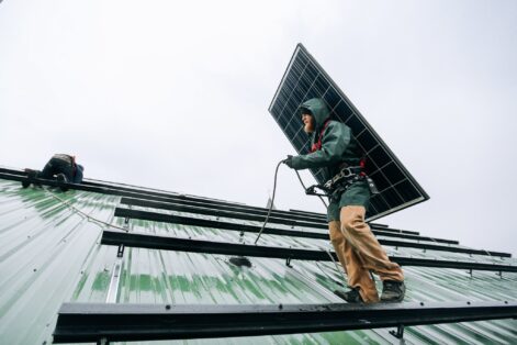 A man stands on a roof of solar panels, carrying another panel, ready to install it