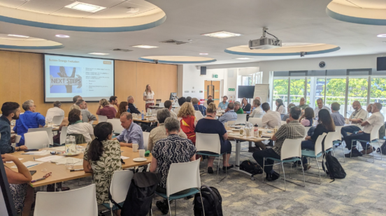 A presentation taking place in a large room with delegates at tables and a speaker at the front with a projector screen